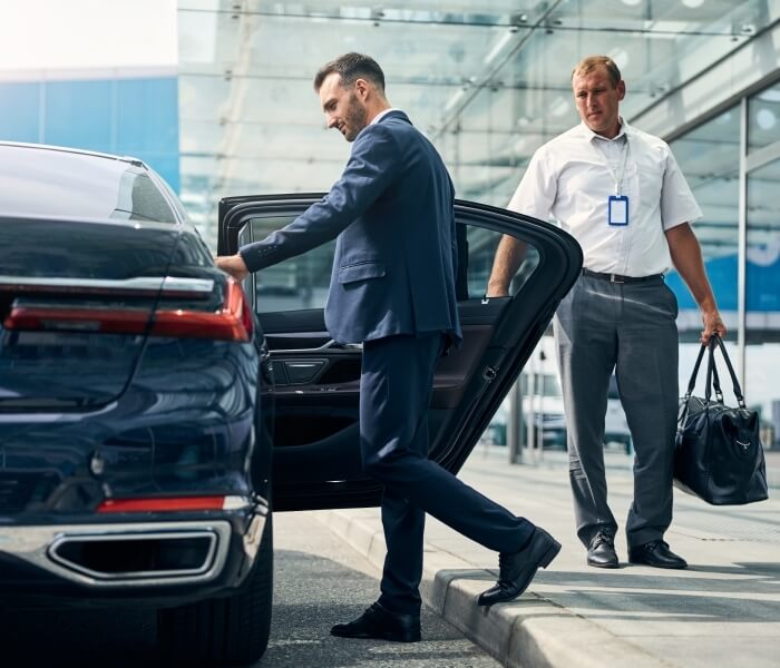 A man in a dark suit exits a car while another man in a white shirt and gray pants stands behind him, holding bags.