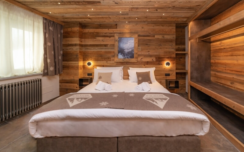 Cozy hotel room with a central bed, wooden accents, lamps, and an open window, evoking a mountain lodge atmosphere.