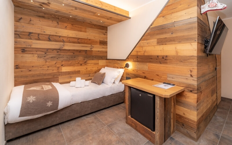 Cozy room with a bed, desk, and wall-mounted TV, featuring light wood paneling and a brown-tiled floor.