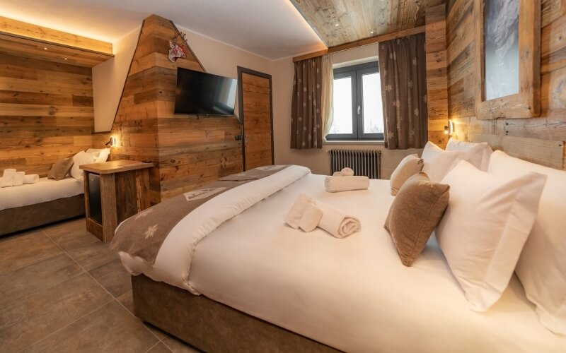 Cozy bedroom with two beds, light wood walls, a TV, window, brown tiled floor, and a small table with a lamp.