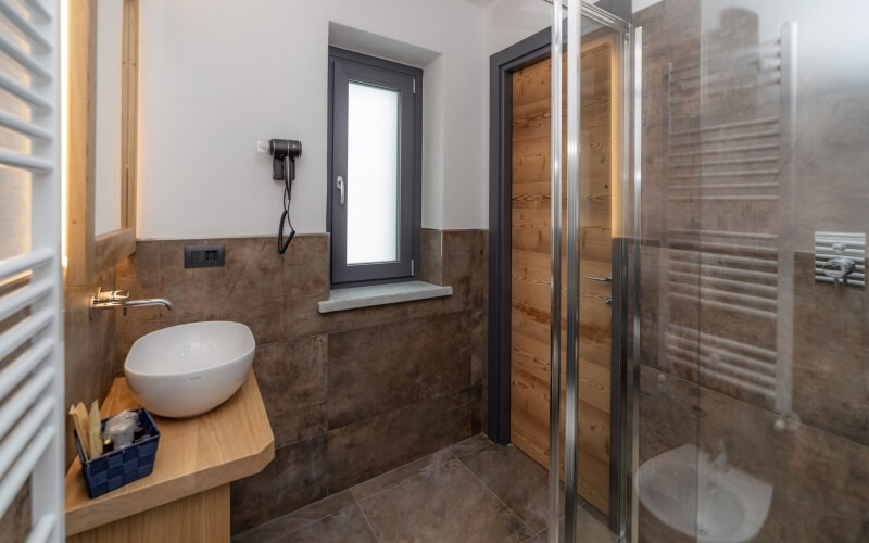 Modern bathroom with wooden sink, white bowl, glass shower, brown tiled walls, and gray tiled floor, exuding simplicity and cleanliness.