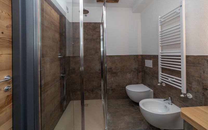 Modern bathroom with glass shower, bidet, towel rack, brown tiles, skylight, and wooden cabinet.