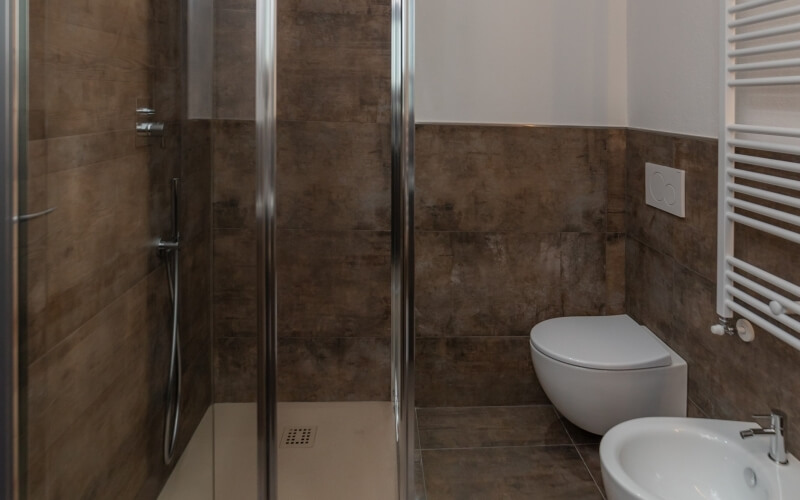 Modern bathroom featuring a glass-panel shower, white toilet, bidet, and dark brown tiles against white walls.