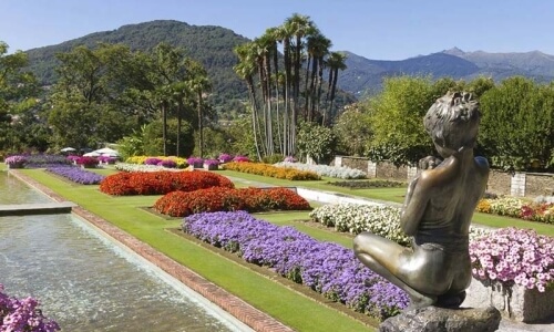 Serene garden with a pond, child statue, colorful flowers, palm trees, and mountains under a blue sky, evoking peace.