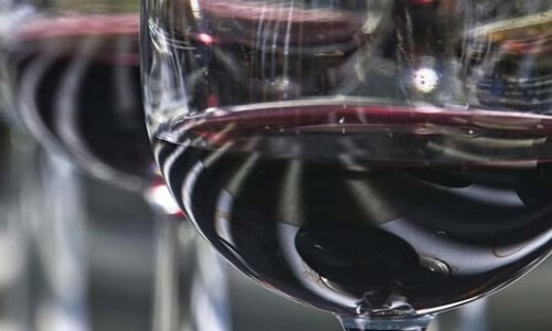 Close-up of a clear wine glass with dark red liquid, reflecting light, against a blurred background of other glasses.