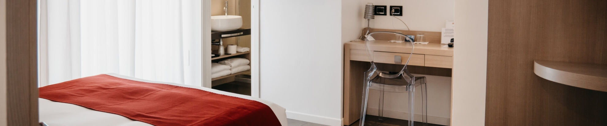 Camera d'hotel con scrivania in legno, letto con coperta rossa, e bagno con lavabo e asciugamani visibili.