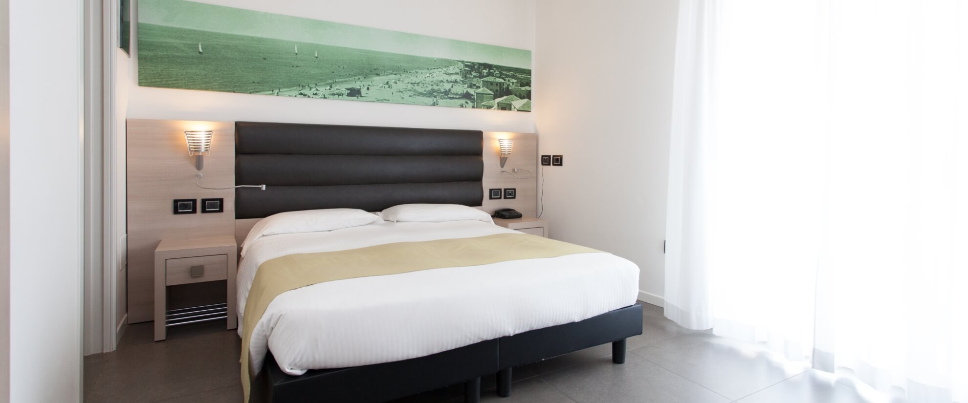 Camera d'hotel moderna con letto grande, comodino, lampada, arte murale e pavimento in piastrelle grigie.
