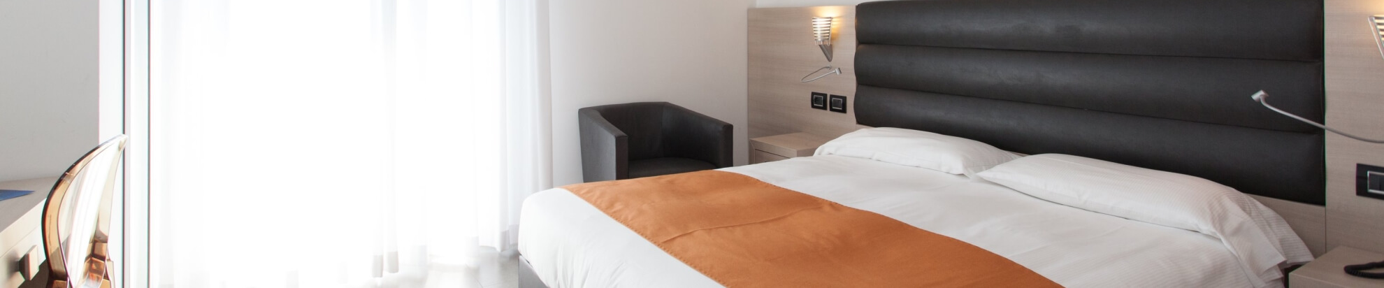 Camera d'albergo moderna con letto bianco, scrivania, finestre con tende bianche e atmosfera minimalista.