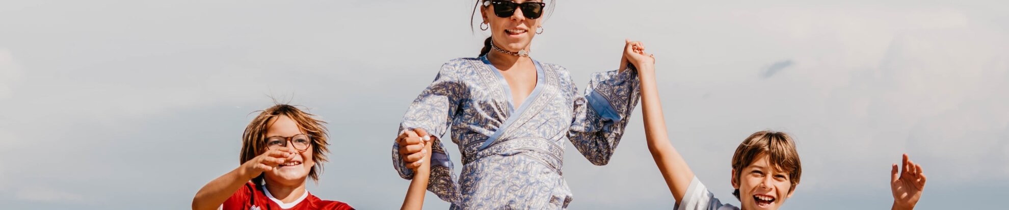 Una donna in abito bianco con fiori blu tiene per mano due bambini sotto un cielo grigio.