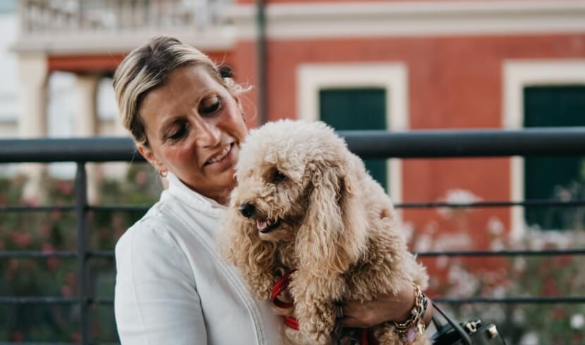 Una donna bionda in giacca bianca tiene un cane marrone chiaro davanti a un parapetto, con un edificio sullo sfondo.