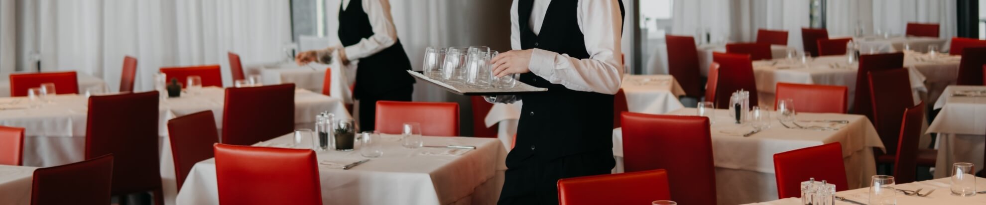 Impostazione di un ristorante elegante con tavoli, sedie rosse e camerieri in abiti formali che servono bicchieri vuoti.
