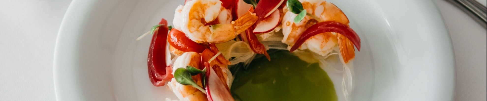 Ciotola bianca con insalata di gamberi, peperoni rossi e foglie verdi su noodles, con brodo verde, su un tavolo bianco.