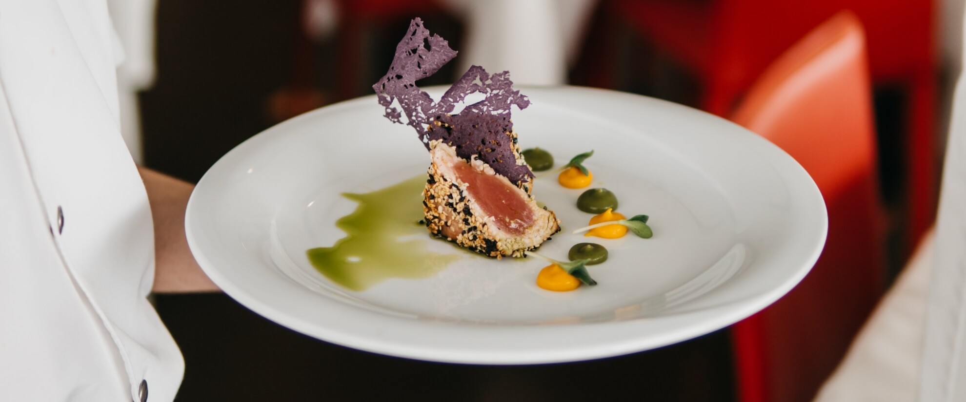 Piatto bianco con carne rosa, semi di sesamo, fagiolini, salsa arancione e guarnizione viola, in un ambiente ristorante.