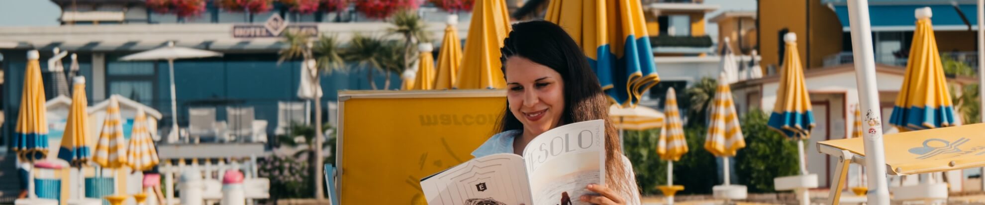 Una donna con capelli scuri legge una rivista "SOLO" su una sedia gialla, con un hotel e palme sullo sfondo.