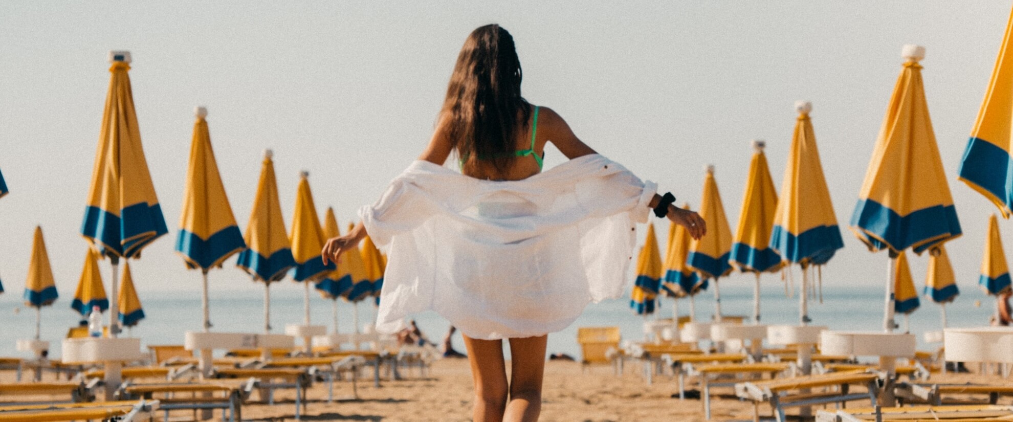 Donna con bikini verde e camicia bianca cammina sulla spiaggia, circondata da ombrelloni gialli e acqua calma.