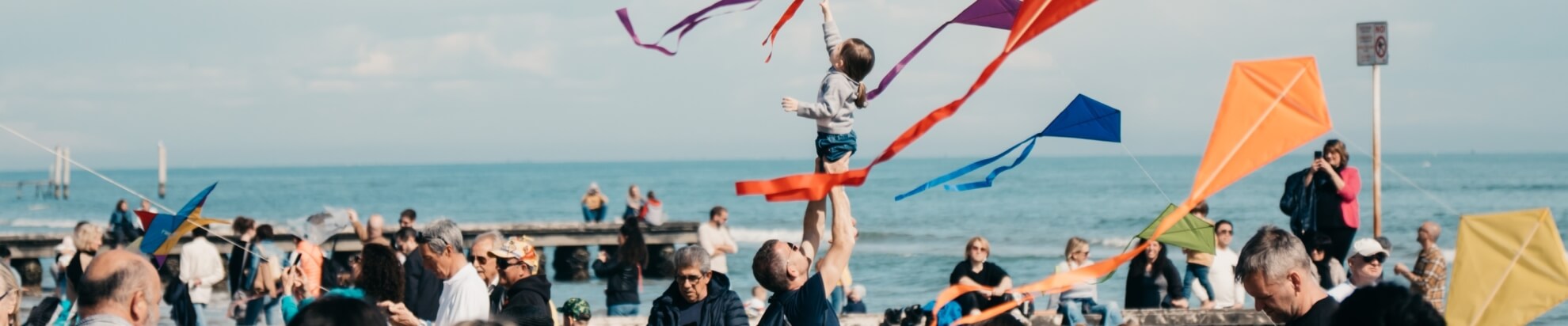 Una scena vivace di persone sulla spiaggia, con un adulto che solleva un bambino e aquiloni colorati che volano nel cielo.