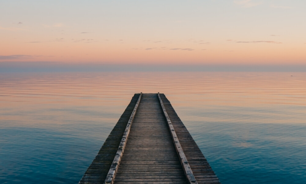 Un molo di legno si estende su acque calme, riflettendo un cielo sereno con sfumature di rosa, arancione e blu.