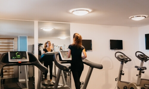 Una donna corre su un tapis roulant in palestra, con un'altra donna e attrezzature visibili sullo sfondo.