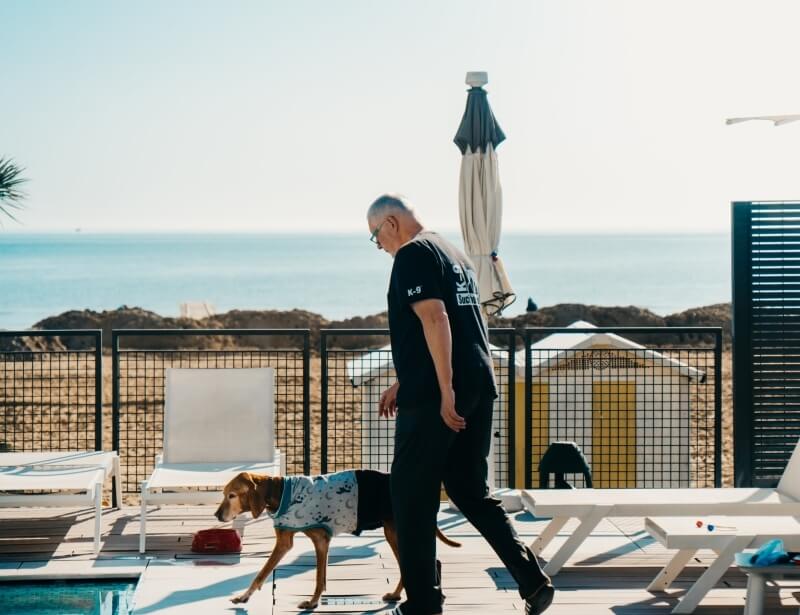 Un uomo in pantaloni neri e t-shirt nera cammina con un cane su un ponte di legno, con l'oceano sullo sfondo.