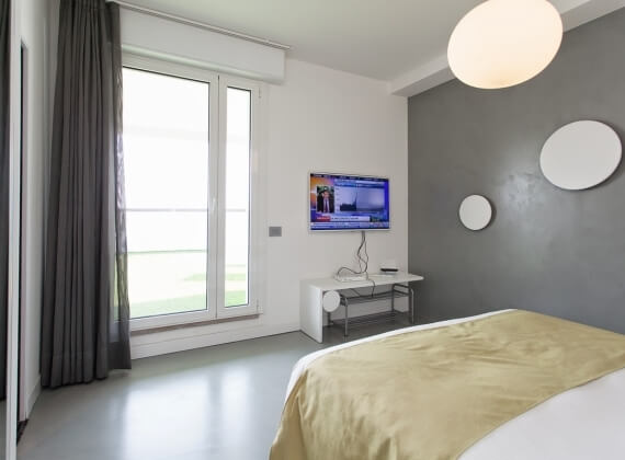 Camera da letto moderna e minimalista con pareti bianche, letto con coperta gialla e grande finestra con tende grigie.