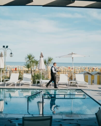 Uomo in giacca che passeggia accanto a una piscina circondata da lettini e ombrelloni, con vista sulla spiaggia.