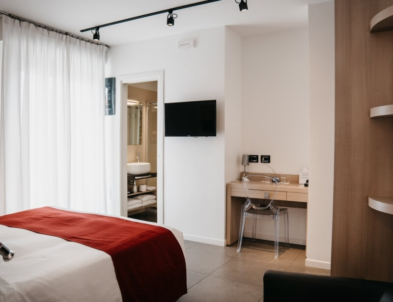 Camera d'albergo moderna con letto, scrivania, bagno e pavimento in piastrelle chiare. Tende bianche e coperta rossa.