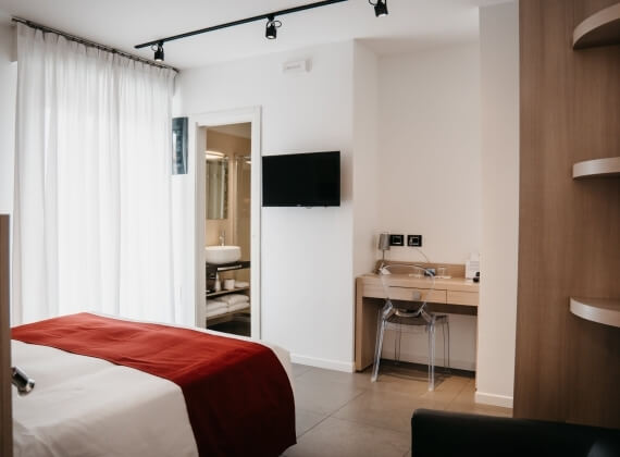 Ein modernes Hotelzimmer mit einem weißen Bett und roter Decke, Schreibtisch, Badezimmer und viel natürlichem Licht.