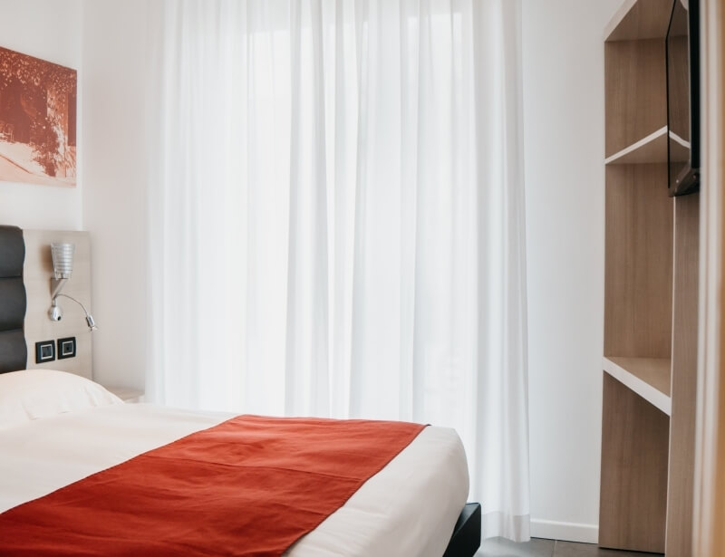 Camera serena con letto bianco e runner rosso, lampada a muro, opere d'arte e tende bianche che coprono una finestra.
