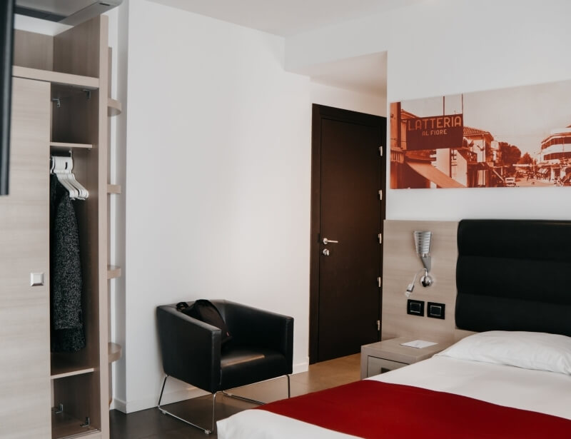 Camera d'albergo con letto, sedia in pelle nera, armadio aperto e foto sepia sopra il letto. Pareti bianche e pavimento in legno chiaro