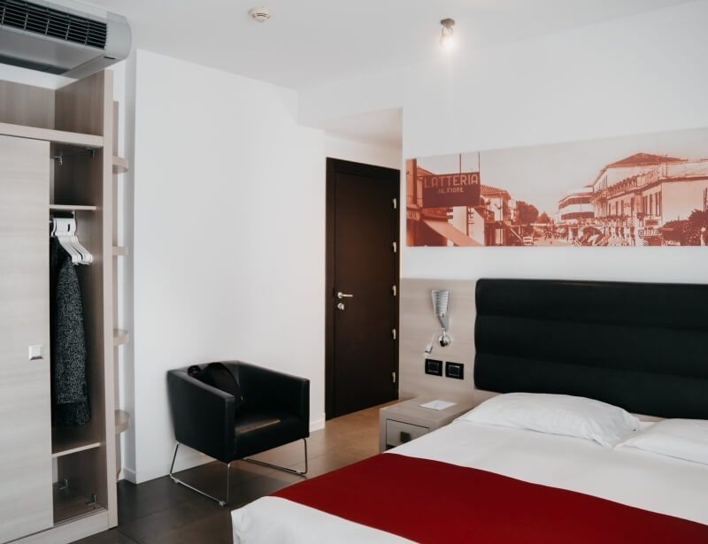 Camera d'albergo moderna e minimalista con letto grande, coperta rossa, poltrona nera e armadio in legno. Atmosfera elegante.