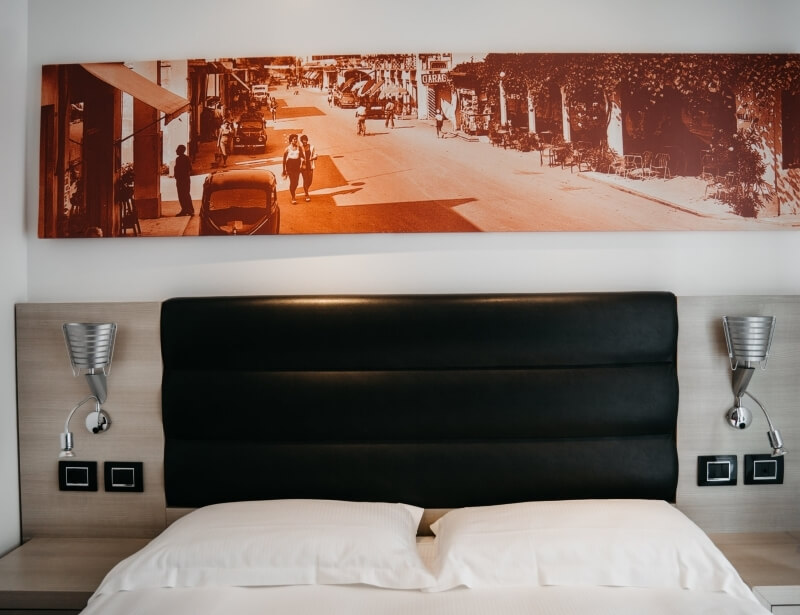 Camera d'hotel con letto, testiera in pelle nera, lampade a muro e fotografia vintage sopra il letto. Atmosfera accogliente.