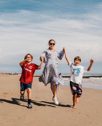 Donna e due ragazzi saltano sulla spiaggia, con il mare sullo sfondo.