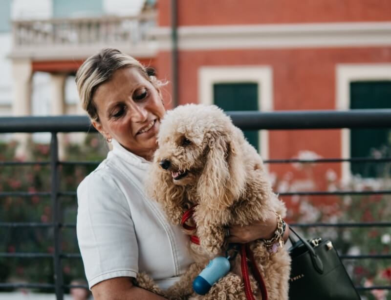 Donna bionda con una maglietta bianca tiene in braccio un cane meticcio, davanti a un edificio rosso con alberi sullo sfondo.