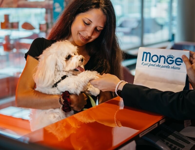 Donna con un cane bianco riceve una borsa "Monge" da un'altra persona al bancone di un negozio per animali.