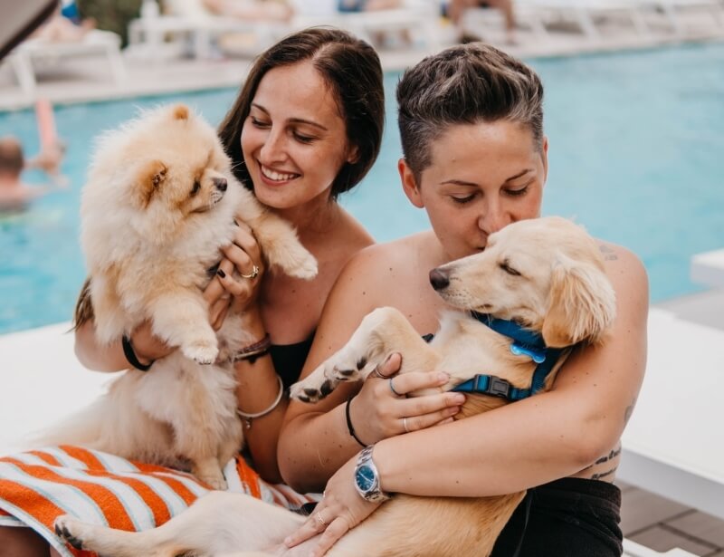 Due donne sedute vicino a una piscina, ciascuna con un cane in braccio, in un'atmosfera serena e rilassata.