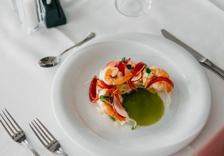 Un piatto bianco con salsa verde, gamberi, peperoni rossi e microgreens, posizionato su una tovaglia bianca elegante.