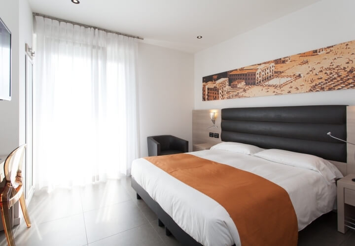 Camera d'hotel con letto matrimoniale, tonalità bianche, grigie e arancioni, e dipinto sopra il letto.