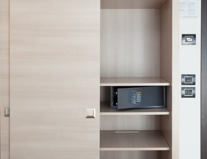 Ein helles Holzschrank mit offener Tür, einem grauen Safe auf dem zweiten Regal und Steuergeräten an der Wand.