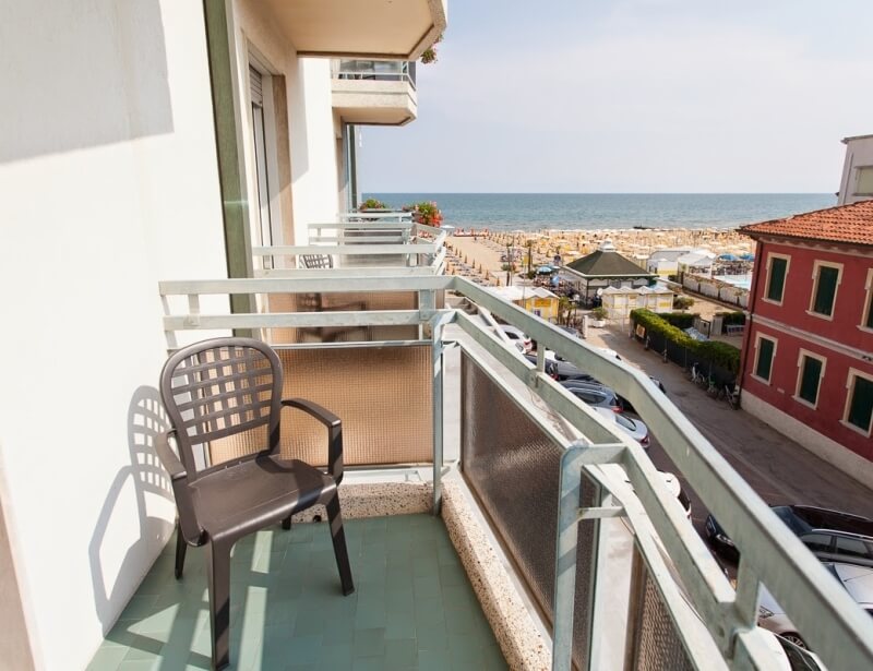Ein ruhiger Strandblick von einem Balkon mit einem Stuhl, weißen Geländern und einem grünen Boden, unter einem blauen Himmel.