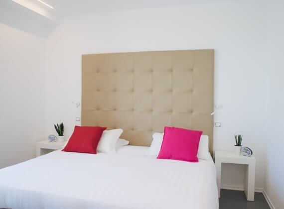 Camera da letto minimalista con testiera beige, letto bianco, cuscini rossi e rosa, piante e luce naturale.