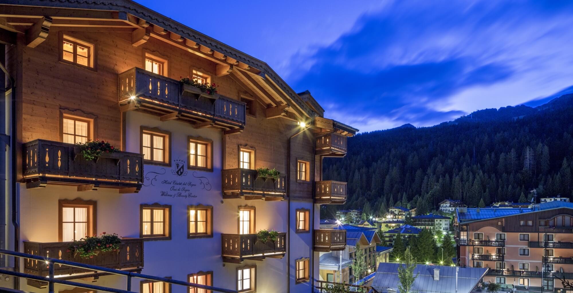 Mehrstöckiges Gebäude mit Balkonen und beleuchteten Fenstern, umgeben von Bergen und Bäumen im Zwielicht.