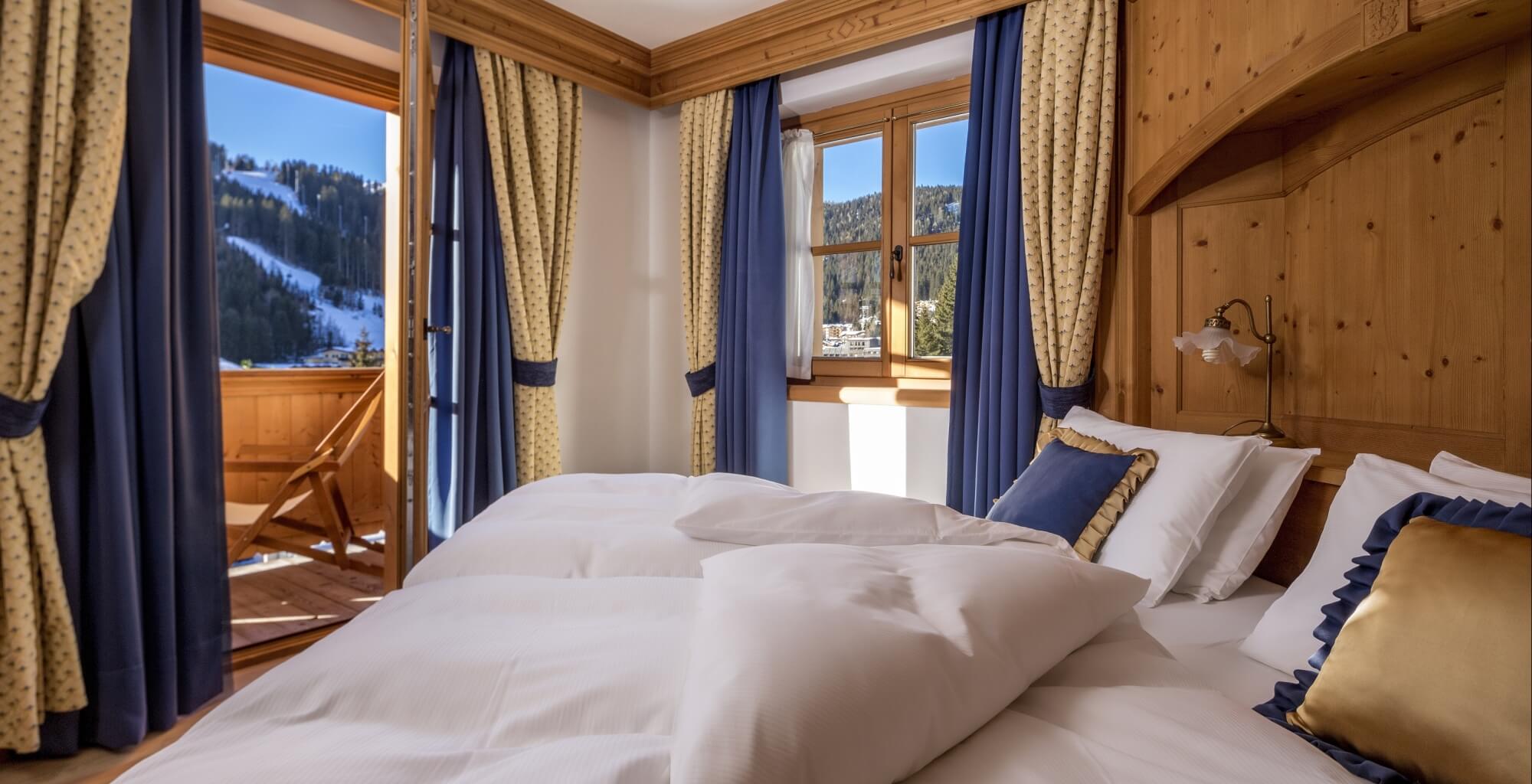 Schlafzimmer mit weißen Bettdecken, Holzkopfteil und Blick auf schneebedeckte Berge durch Fenster. Antike Lampe am Kopfteil.