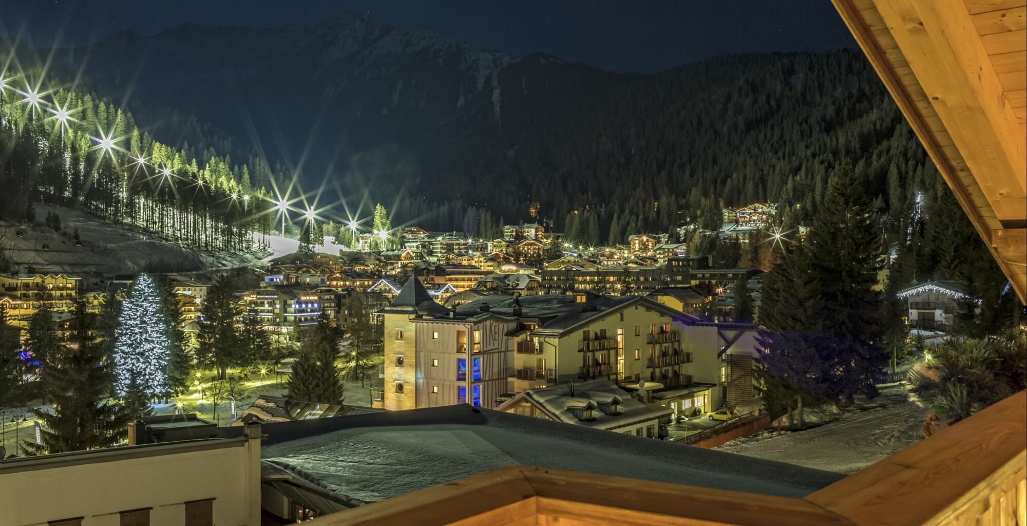 Nachtaufnahme eines beleuchteten Bergdorfes im Schnee, umgeben von Tannen, mit einem großen Weihnachtsbaum in der Mitte.