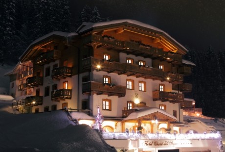 Edificio bianco a più piani con balconi illuminati, situato su una collina innevata, circondato da alberi scuri.