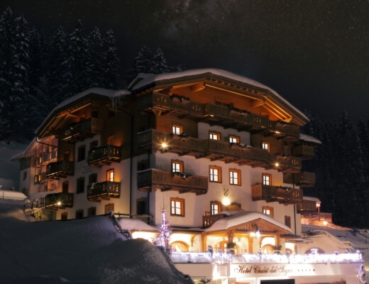 Edificio multi-storico bianco con accenti marroni, illuminato, circondato da alberi innevati e cielo stellato.