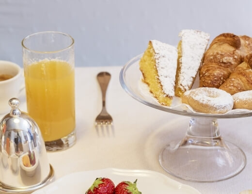 Breakfast table with orange juice, coffee mug, strawberries, and a cake stand with croissants and pastries against a white wall.