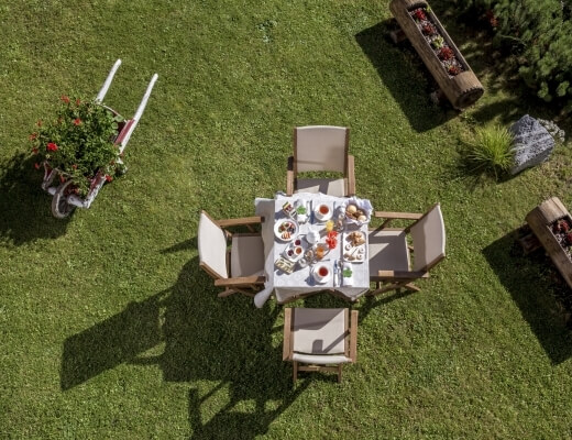 A serene outdoor setting with a table and chairs on a green lawn, adorned with dishes and surrounded by potted plants.