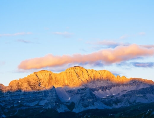 Catena montuosa illuminata dal sole con cielo blu e nuvole sparse, tonalità di blu e arancione sui picchi.