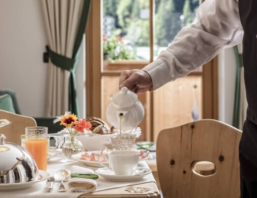 A person pours tea into a cup at a cozy breakfast table with food, flowers, and a view of trees through a window.