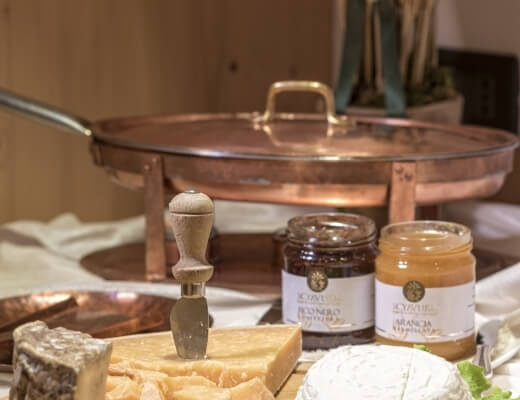 Table with jars of jam, cheese, knife, and a copper pan, set against a wooden wall and white cloth.